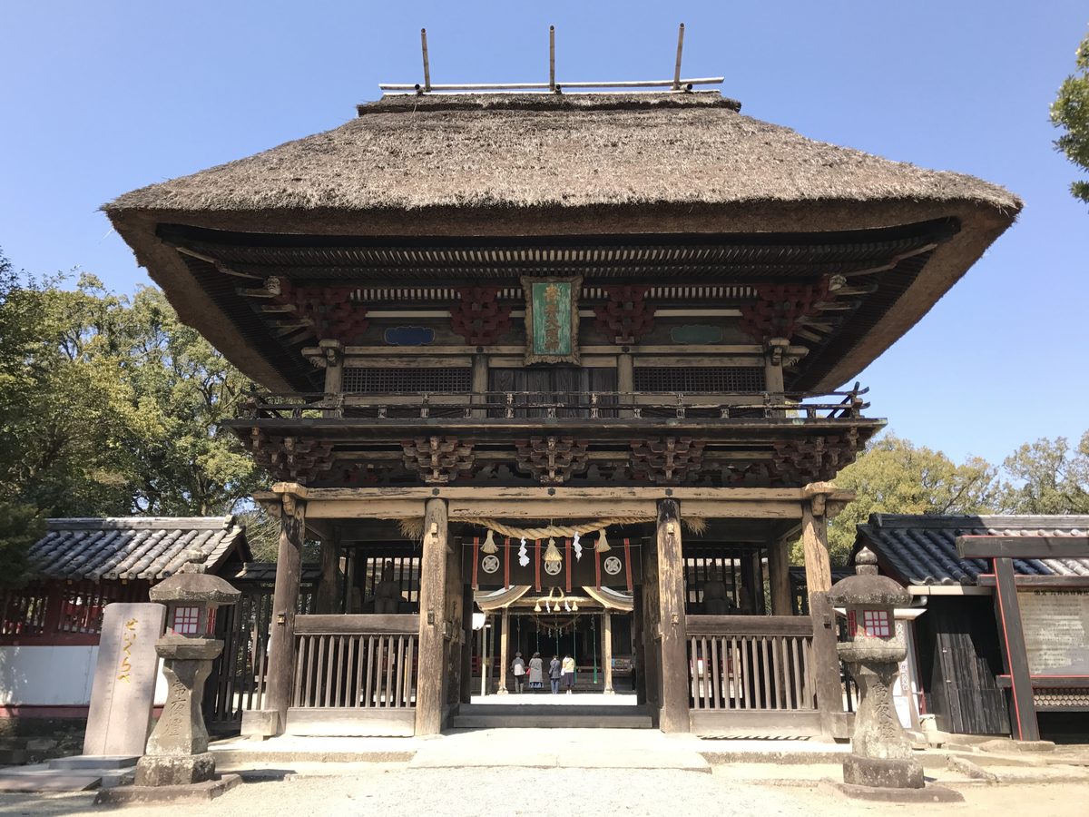 The two-storied Romon gate of Aoi Aso Shrine in Hitoyoshi, with a steep thatched roof, carved red and black details and stone lanterns on each side.