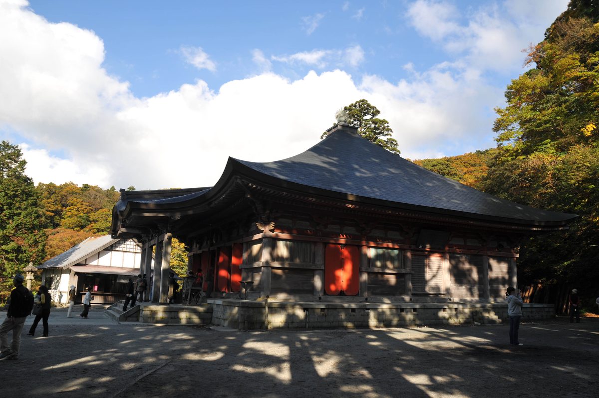 The main hall of Daisen-ji Temple, a wide wooden hall with a dark curved roof and vermilion doors, with autumn trees on the slope behind and visitors on the gravel ground.