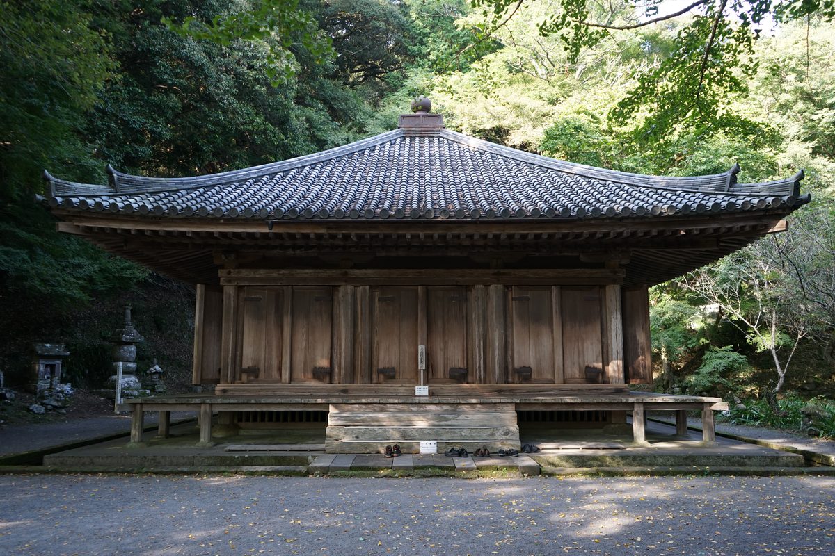 A square wooden hall with a pyramid-shaped tiled roof and plain wooden doors, standing on a low stone base in front of woods.