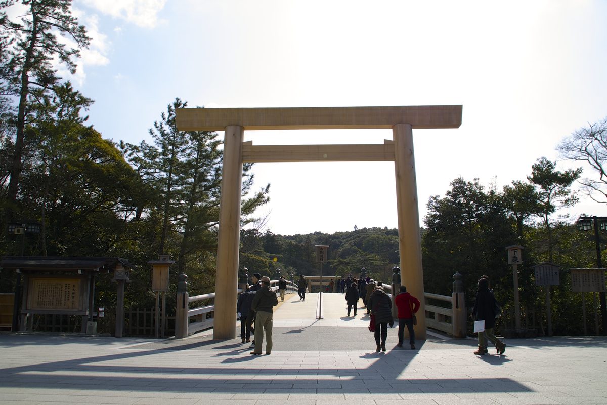 A large plain wooden torii at the end of a bridge, with visitors walking toward it.