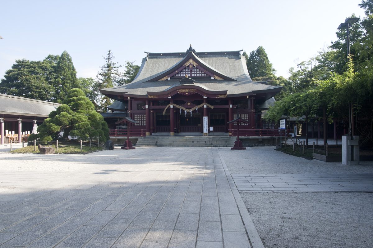The worship hall of Kasama Inari Shrine, painted red with a grey curved roof, seen from the front across a wide stone-paved courtyard, with a pine tree on the left.