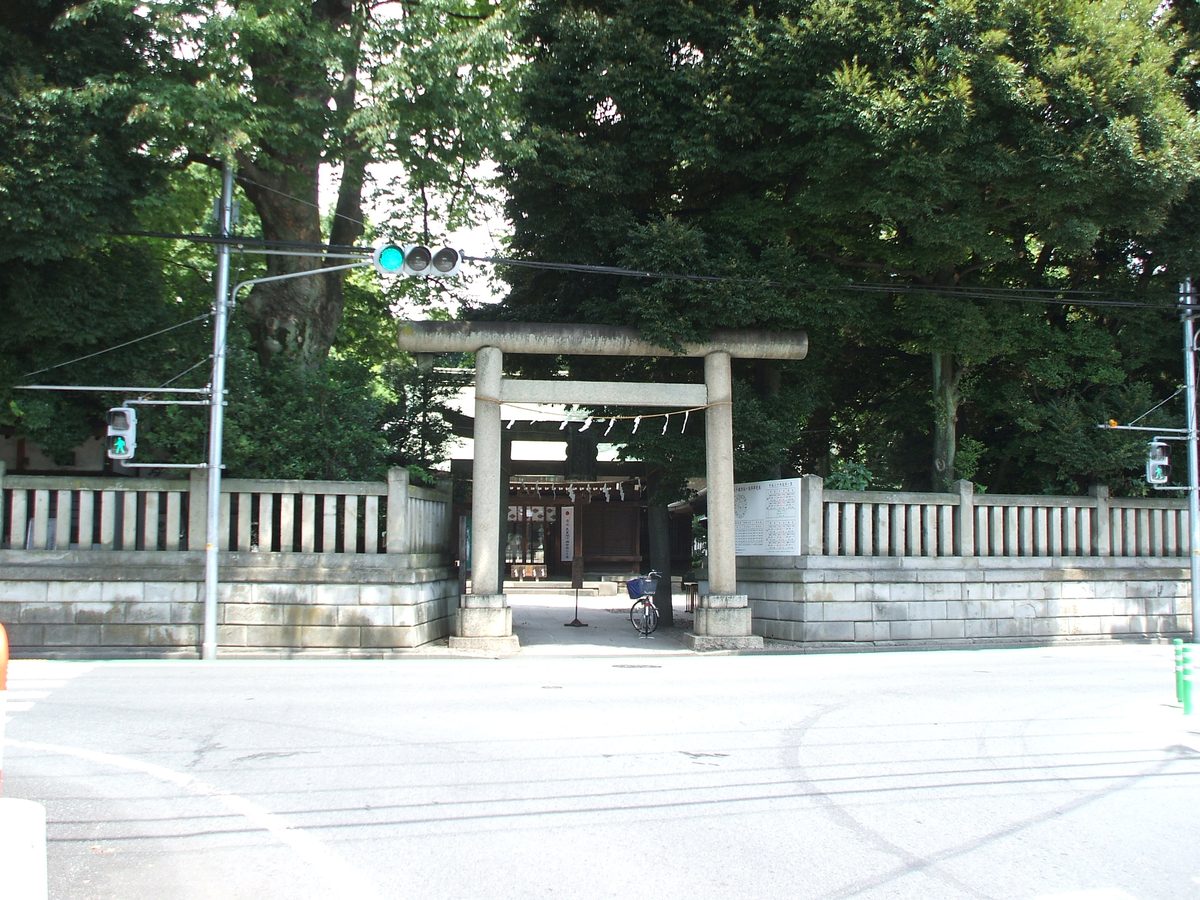 A stone torii gate at the entrance to the shrine grounds, set in a stone and wooden fence beside a street, with large trees behind.