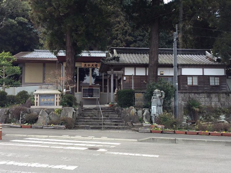A low building with a wooden name board above its entrance, reached by stone steps from the road, with a stone marker on the left and a guardian figure statue on the right.