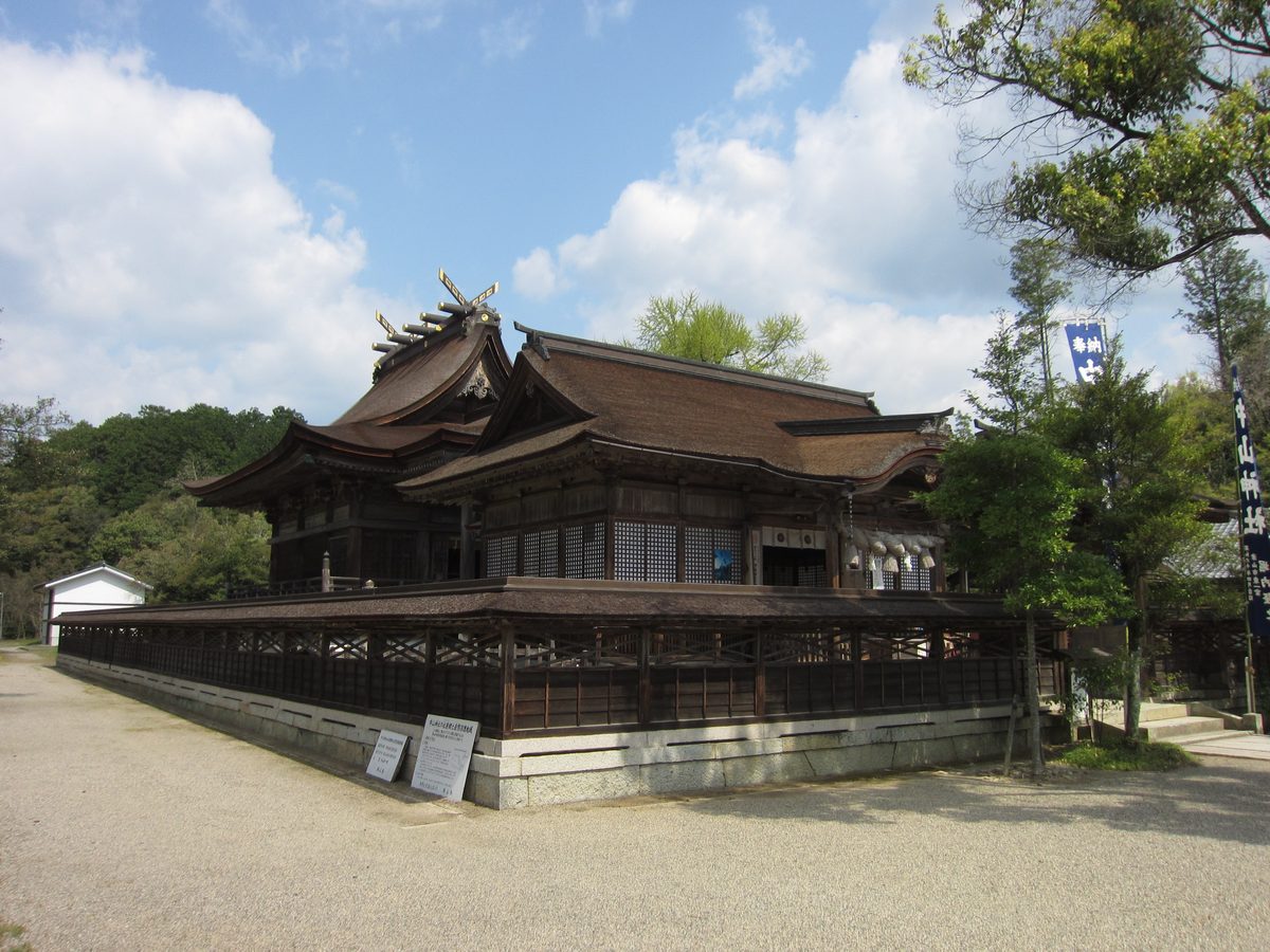 The buildings of Nakayama Shrine seen from an angle, with dark bark-shingled roofs above a low wooden fence, on a gravel ground under a blue sky with clouds.