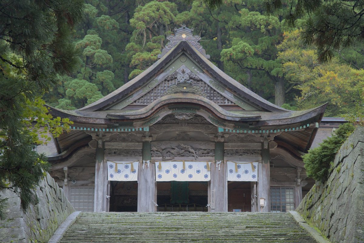 The front of the Okumiya of Ogamiyama Shrine at the top of a flight of stone steps, with a carved gable, white curtains with blue crests and tall cedars behind.