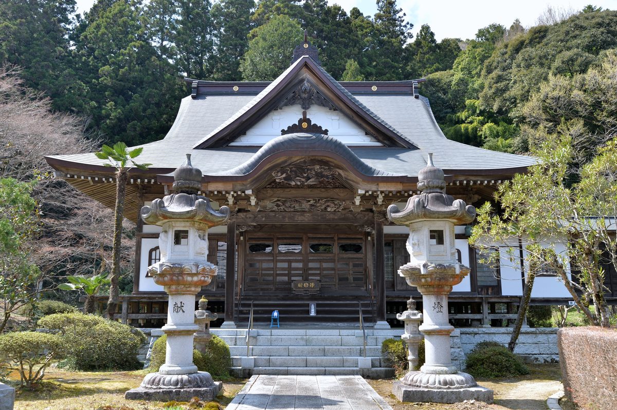 The main hall of Ryogon-ji Temple, with a grey curved roof and a carved wooden front, seen along a stone path between two tall stone lanterns, with wooded hills behind.