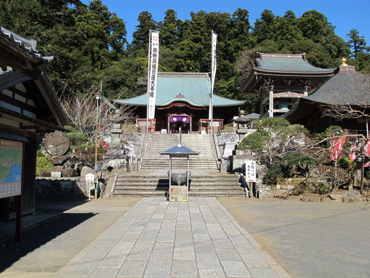 A stone-paved approach and a flight of stone steps leading up to a main hall with a green roof, with a bell tower on the right and wooded hills behind.